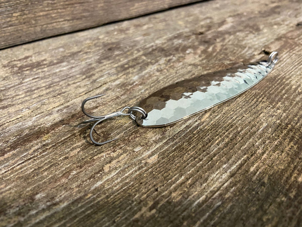 A chatter bass fishing spoon. Two hex style spoons sandwiched together and held together with stainless steel split-rings at the front and back. Also attached, a treble hook, both the lure and hook laying flat on a plank of grey and brown weathered wood.