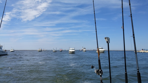 A pack of sportfishing boats following a fishing boat through pier heads with a lighthouse in the background.