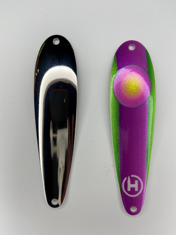 On the right there is a pink, purple, green, and yellow bass fishing lure with a small white HangryBrand logo on it. On the left there is a smooth, shiny, nickel-plated bass fishing spoon.