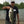 Load image into Gallery viewer, Jeff Spurgess wearing a black t-shirt and cargo shorts holding a large mouth bass with a gravel shoreline and small lake in the background.
