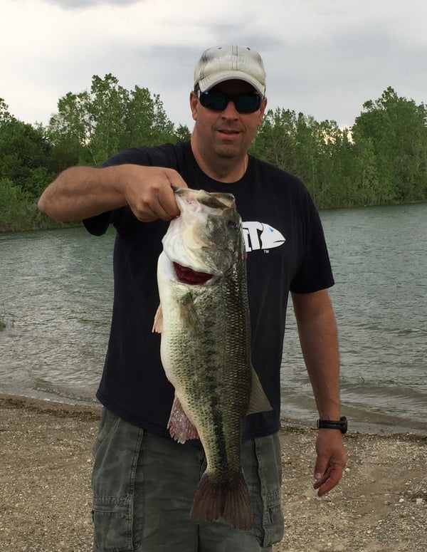 Jeff Spurgess wearing a black t-shirt and cargo shorts holding a large mouth bass with a gravel shoreline and small lake in the background.