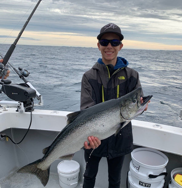 Victor Froman holding a salmon fish while standing in a boat with down riggers and Lake Michigan in the background.