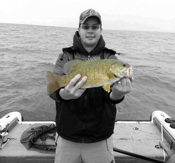 Michael Hiller standing in a Boat holding a small mouth bass. There is a fishing net and choppy water in the background.