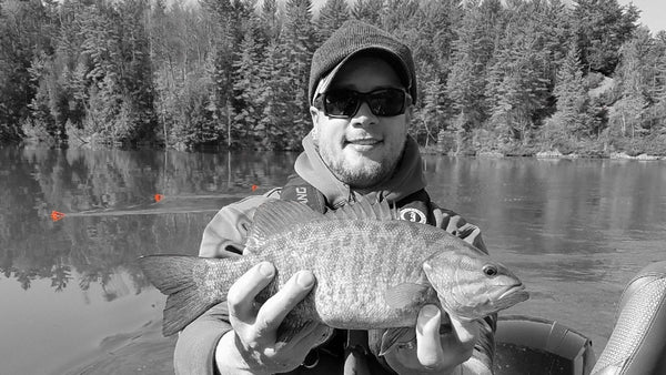 Close up of Michael Hiller wearing a winter hat and standing in a boat holding a small mouth bass. There are three, orange trolling boards in water the background and a line of trees at the edge of a reservoir.