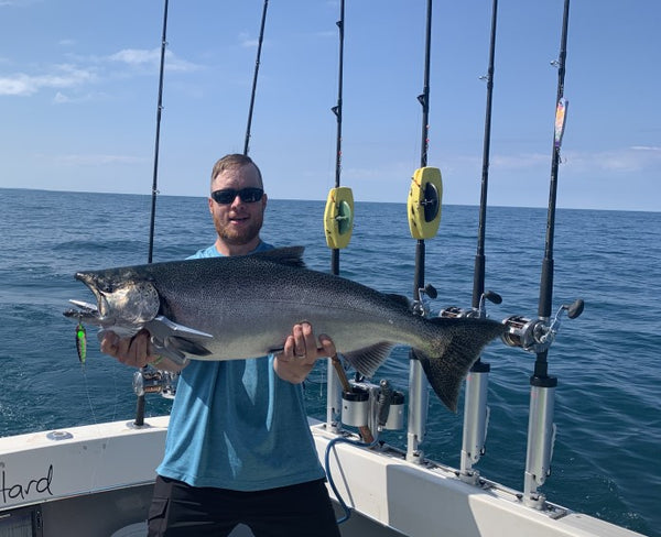 Michael James Hiller standing in the back of a sportfishing boat, sporting a faux-hawk hair cut and wearing sunglasses, and holding a large King Salmon fish with a fishing spoon in the Salmon's mouth.